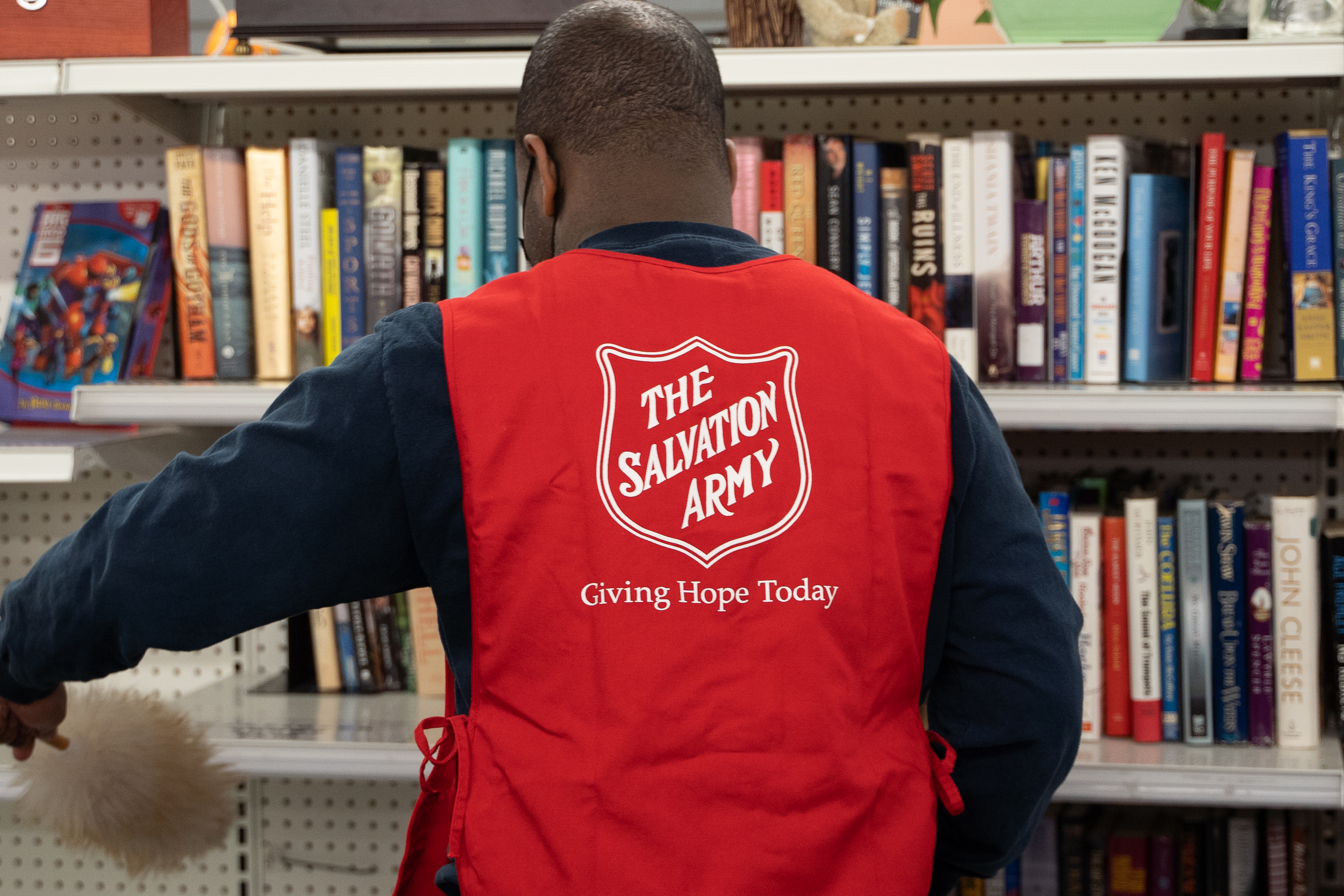 Person wearing a Salvation Army vest organizing books on shelves inside a thrift store.